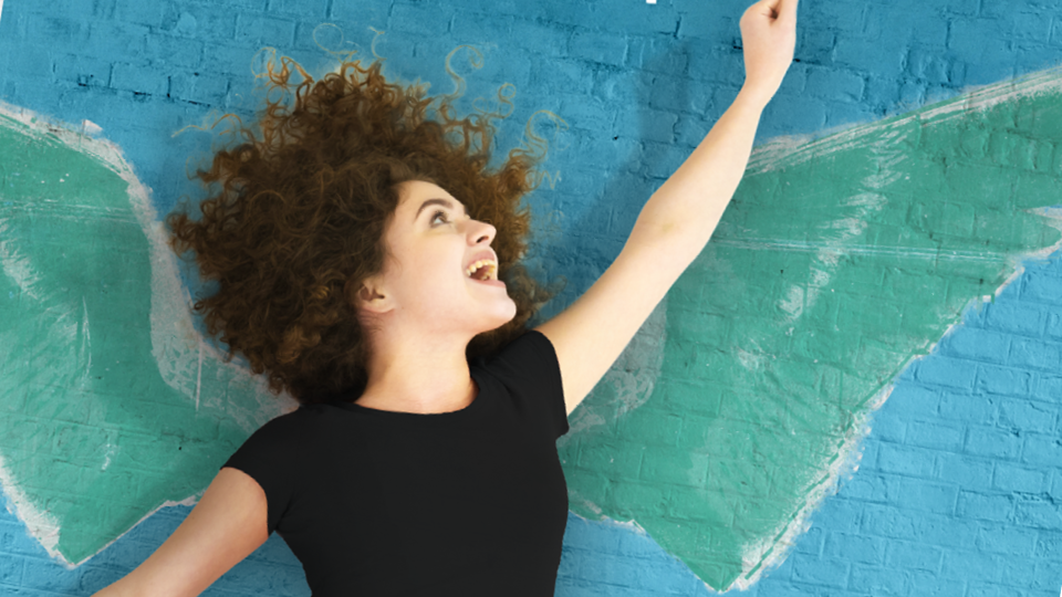 Jeune femme enjouée qui lève le bras au ciel, avec un graffiti d'ailes sur un mur bleu en arrière-plan.